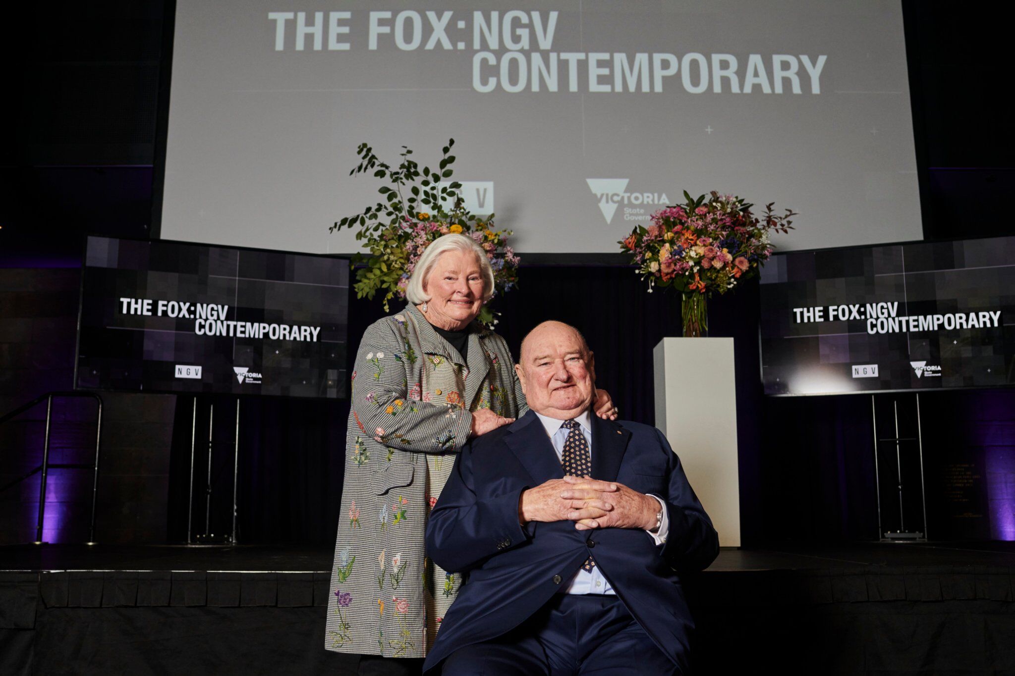 A seated man in a dark grey suit and a woman standing with her hands on the mans shoulder, she is wearing a grey jacket with a floral design. They stand in a dark auditorium with the words The Fox: NGV Contemporary on three screens in the background.