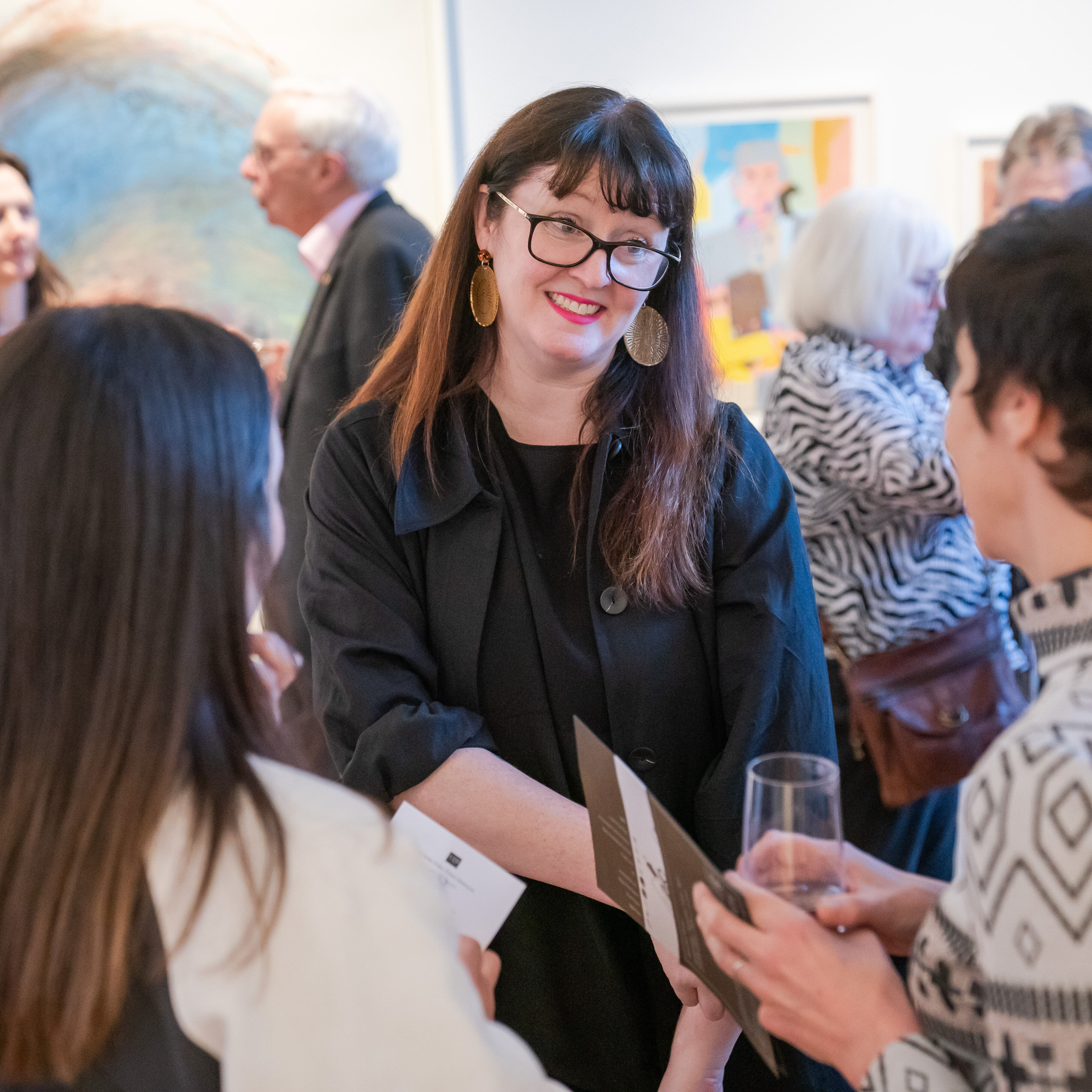 Three women standing and talking to one another, two are pictured from behind and a dark haired woman wearing glasses and a black dress is pictured in the centre. There are people standing in the background and appear out of focus. A number of artworks are on the wall in the background. Photo by James Gifford-Mead