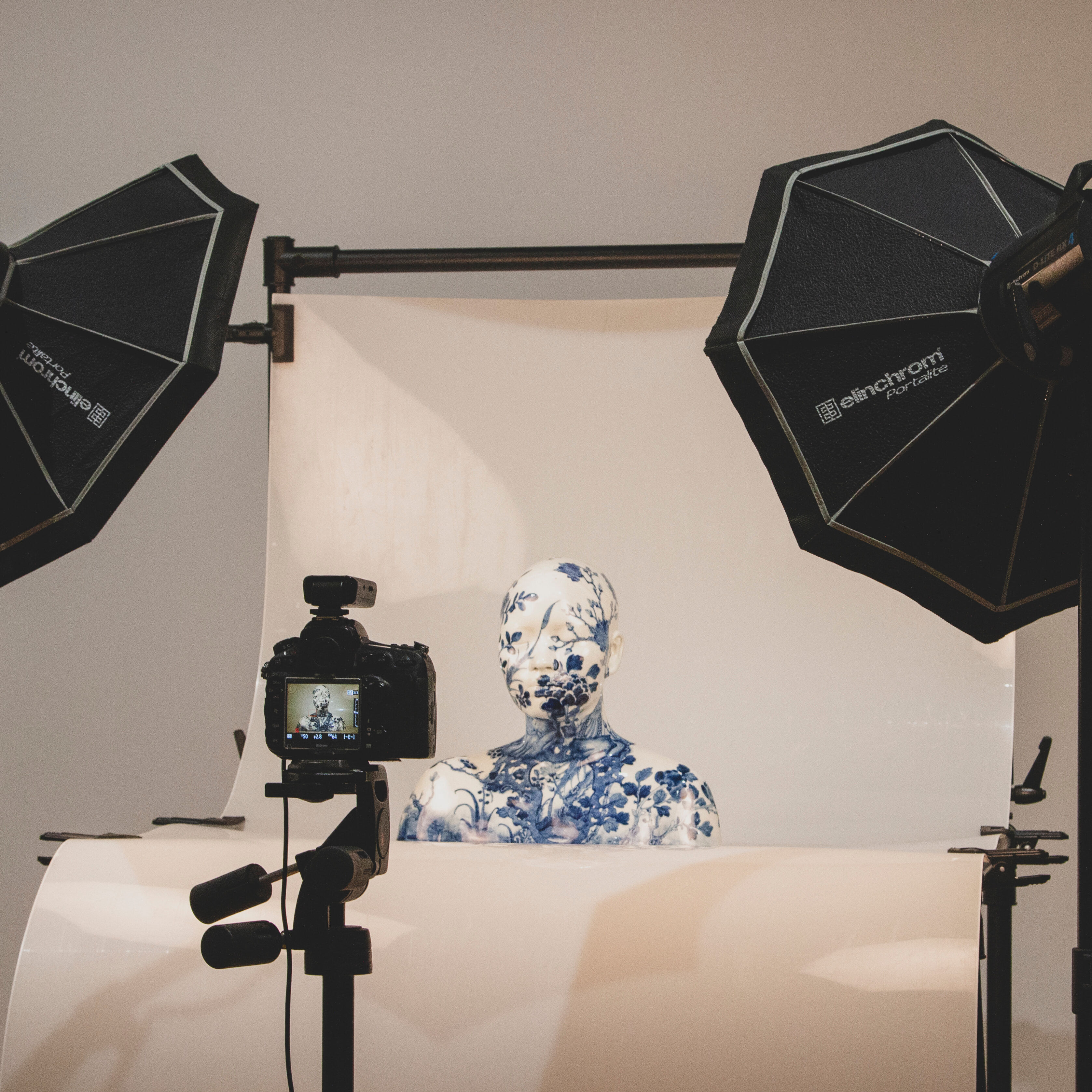 A white ceramic bust with blue floral design overall is sitting against a white photography backdrop, in the foreground is a camera with the bust visible in the viewfinder. Two large black studio lights stand on either side of the ceramic bust.
