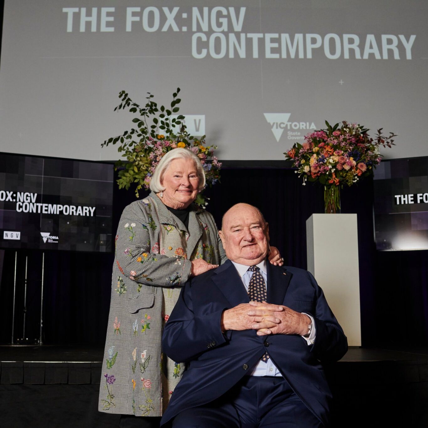 A seated man in a dark grey suit and a woman standing with her hands on the mans shoulder, she is wearing a grey jacket with a floral design. They stand in a dark auditorium with the words The Fox: NGV Contemporary on three screens in the background.