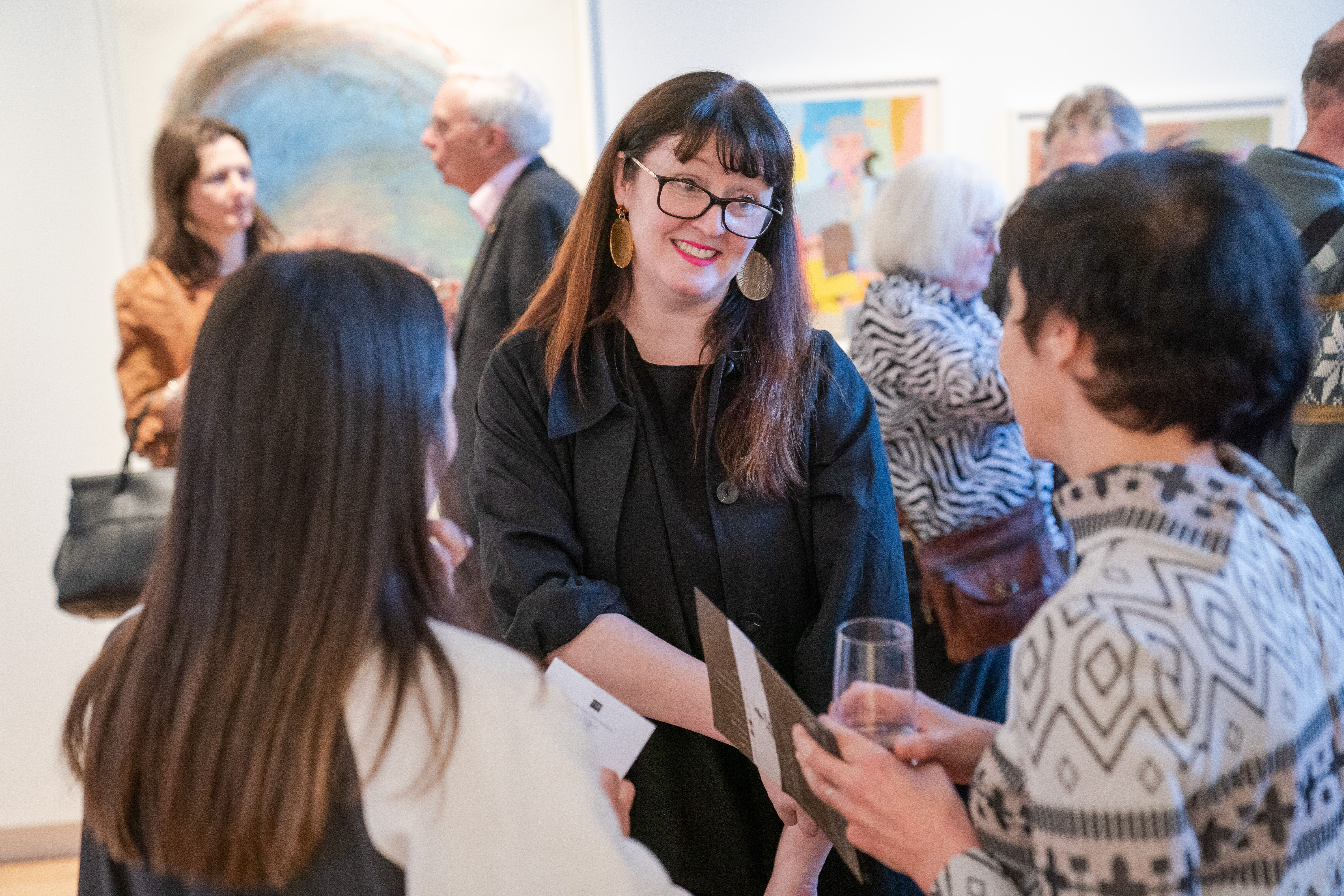 Three women standing and talking to one another, two are pictured from behind and a dark haired woman wearing glasses and a black dress is pictured in the centre. There are people standing in the background and appear out of focus. A number of artworks are on the wall in the background. Photo by James Gifford-Mead