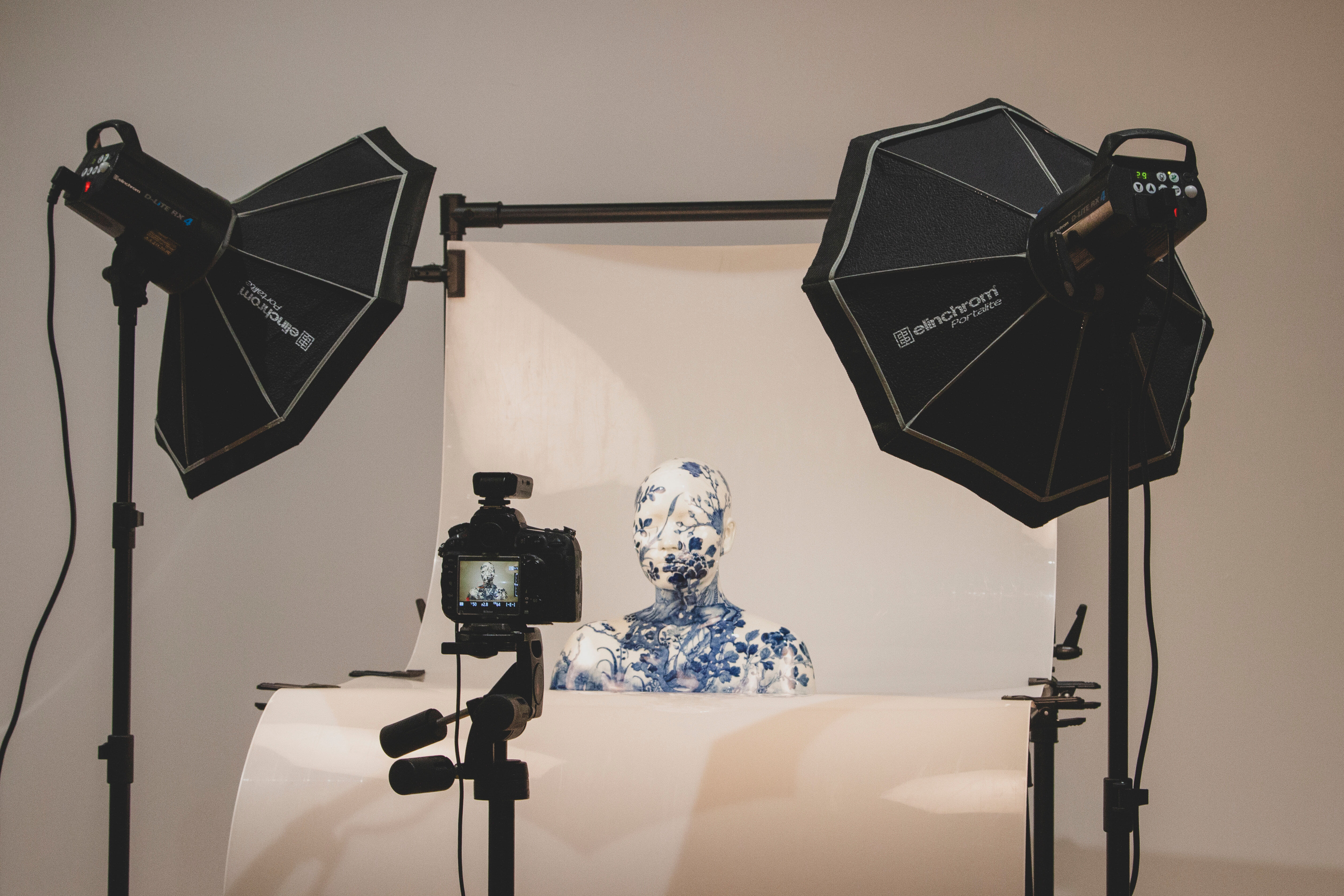 A white ceramic bust with blue floral design overall is sitting against a white photography backdrop, in the foreground is a camera with the bust visible in the viewfinder. Two large black studio lights stand on either side of the ceramic bust.